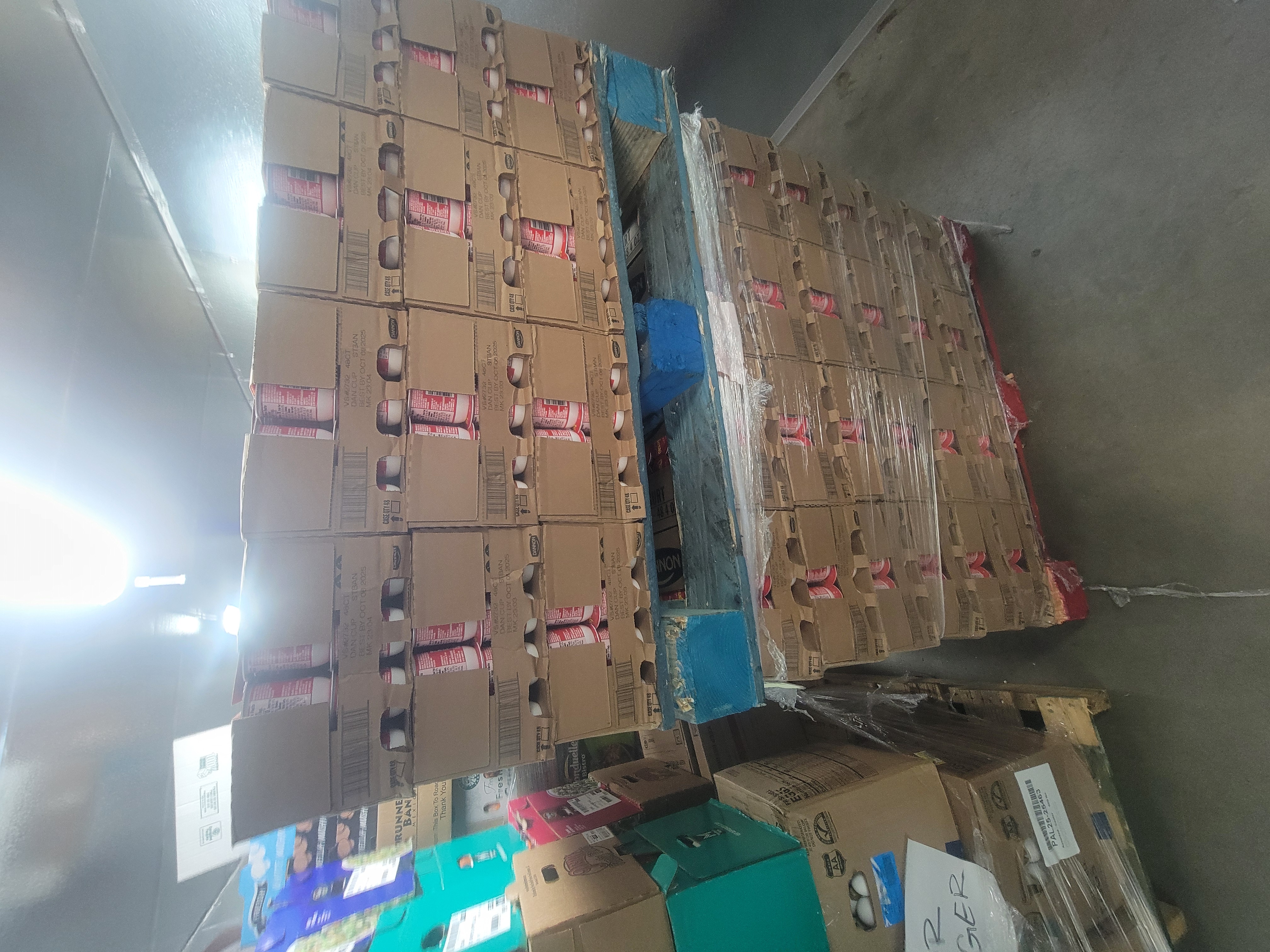 Pallet of cups of yogurt stacked on another pallet of the same thing with a pallet of assorted produce next to it